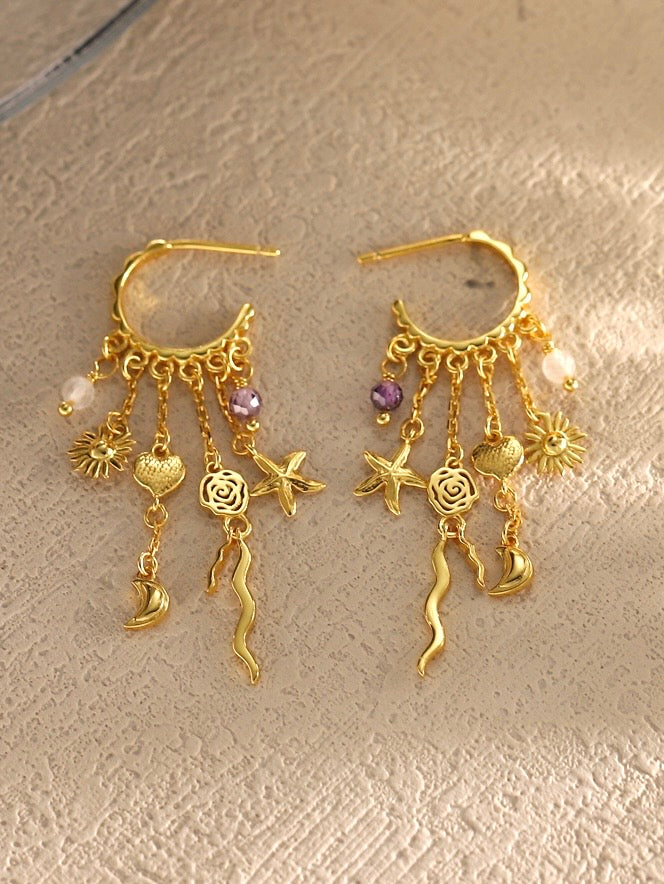 Lilla Havdrømme Øreringe fra Pallantis: Forgyldt sølvøreringe med fem vedhæng hver—søstjerne, rose, hjerte, sol og bølget linje samt lilla og klare perler—præsenteret på hvid baggrund.