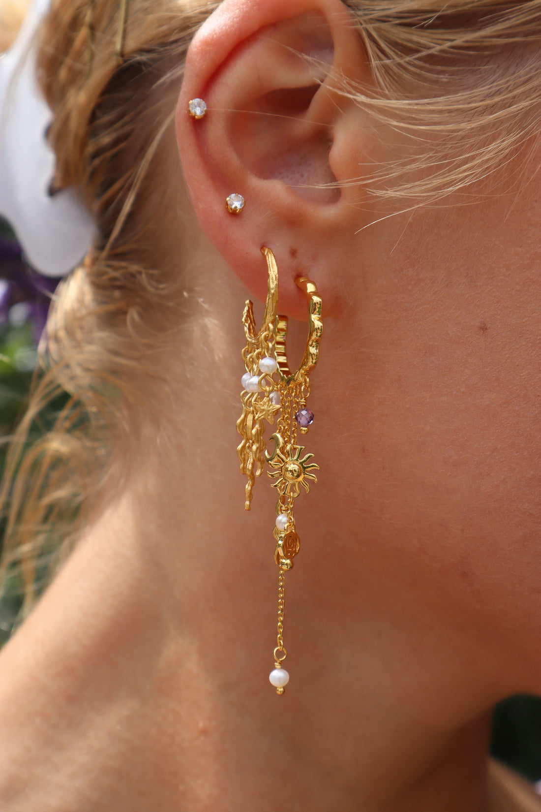 Lilla Sol Øreringe fra Pallantis Smykker: Forgyldte sølvringe med bølgede kanter med dinglende vedhæng – sole, måner, perler, lilla perler, bølgede linjer og blomstercirkler – vist på en hvid baggrund.