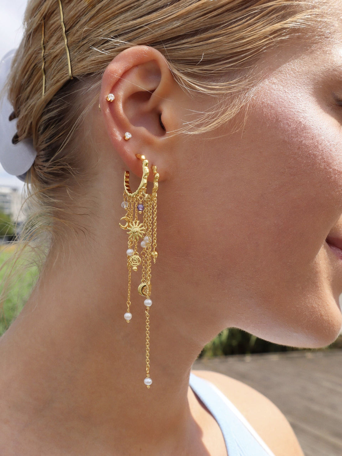 Pallantis Månestjerne Øreringe er bølgede bøjleøreringe i guld med dinglende kæder med charme fra sol, stjerne og halvmåne samt hvide perler – ideel guldøreringe for elskere af himmelske smykker.