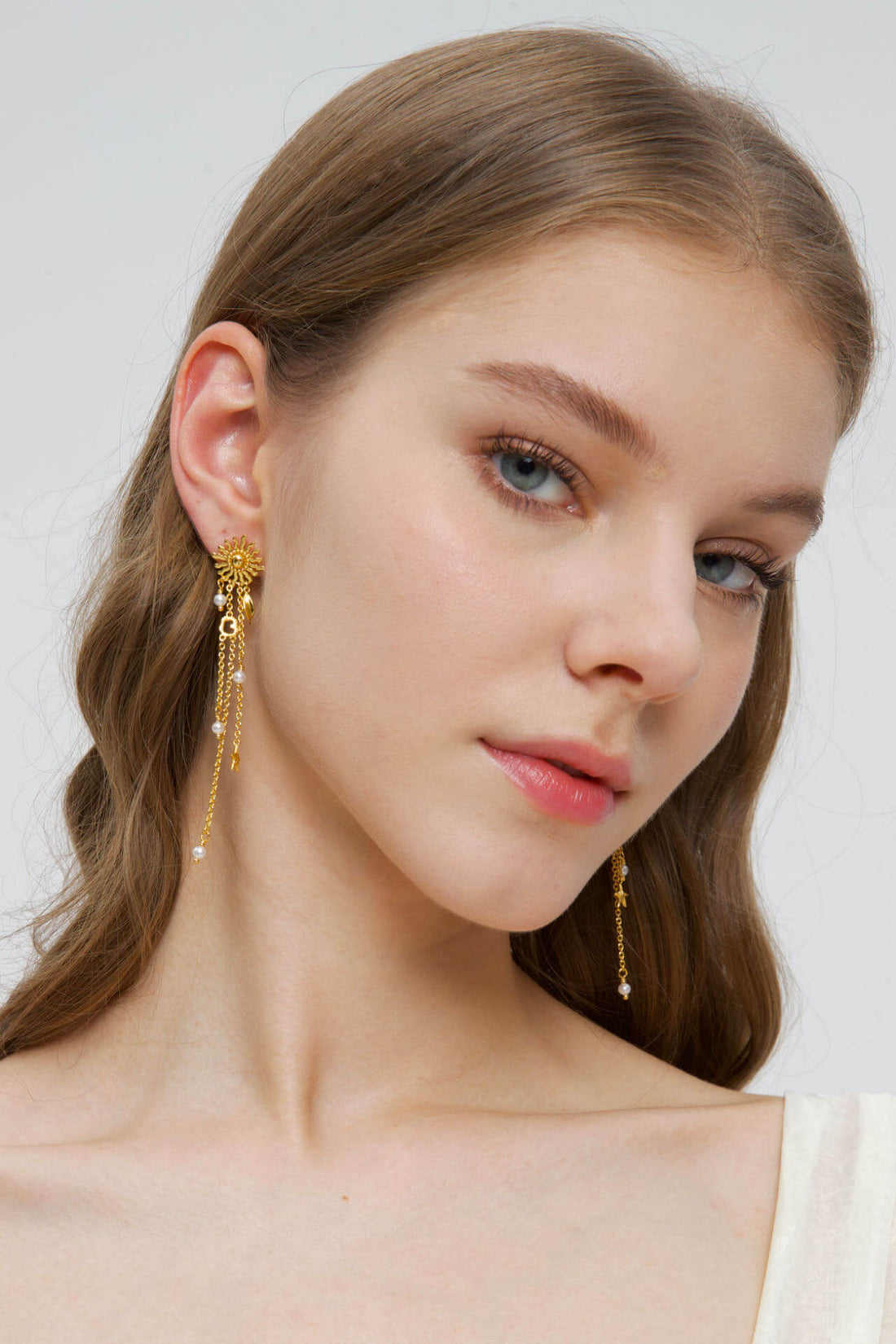 Et par lange, gyldne Sollys-øreringe fra Pallantis Smykker med solformede toppe, dinglende kæder med små guldvedhæng, ferskvandsperler og bladdetaljer.