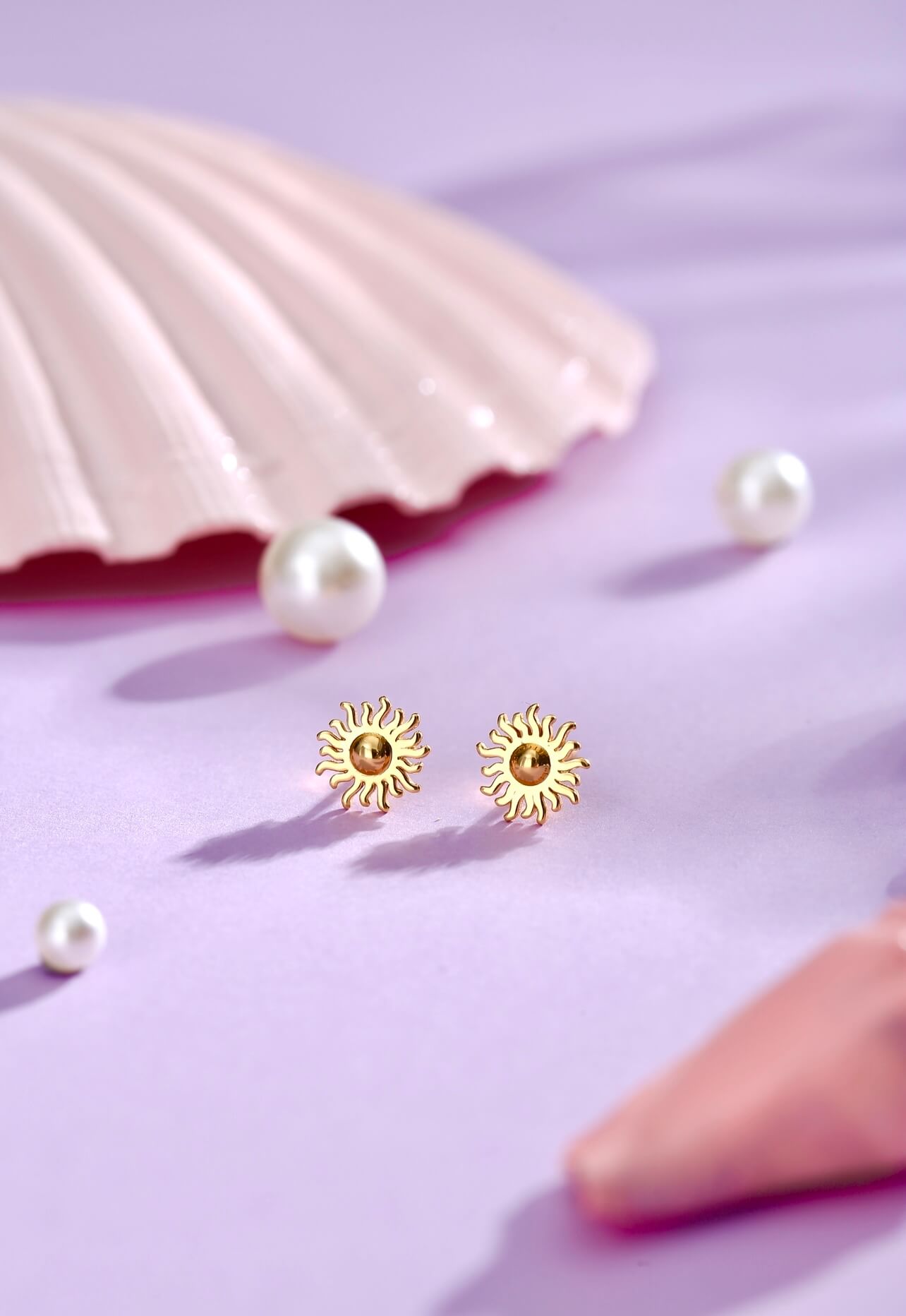 Vivian Sol Ørestikker fra Pallantis Smykker i forgyldt sølv, formet som små sole, vises på en lavendelfarvet overflade blandt perler, med et pink muslingeskal og en geometrisk pink genstand i baggrunden.