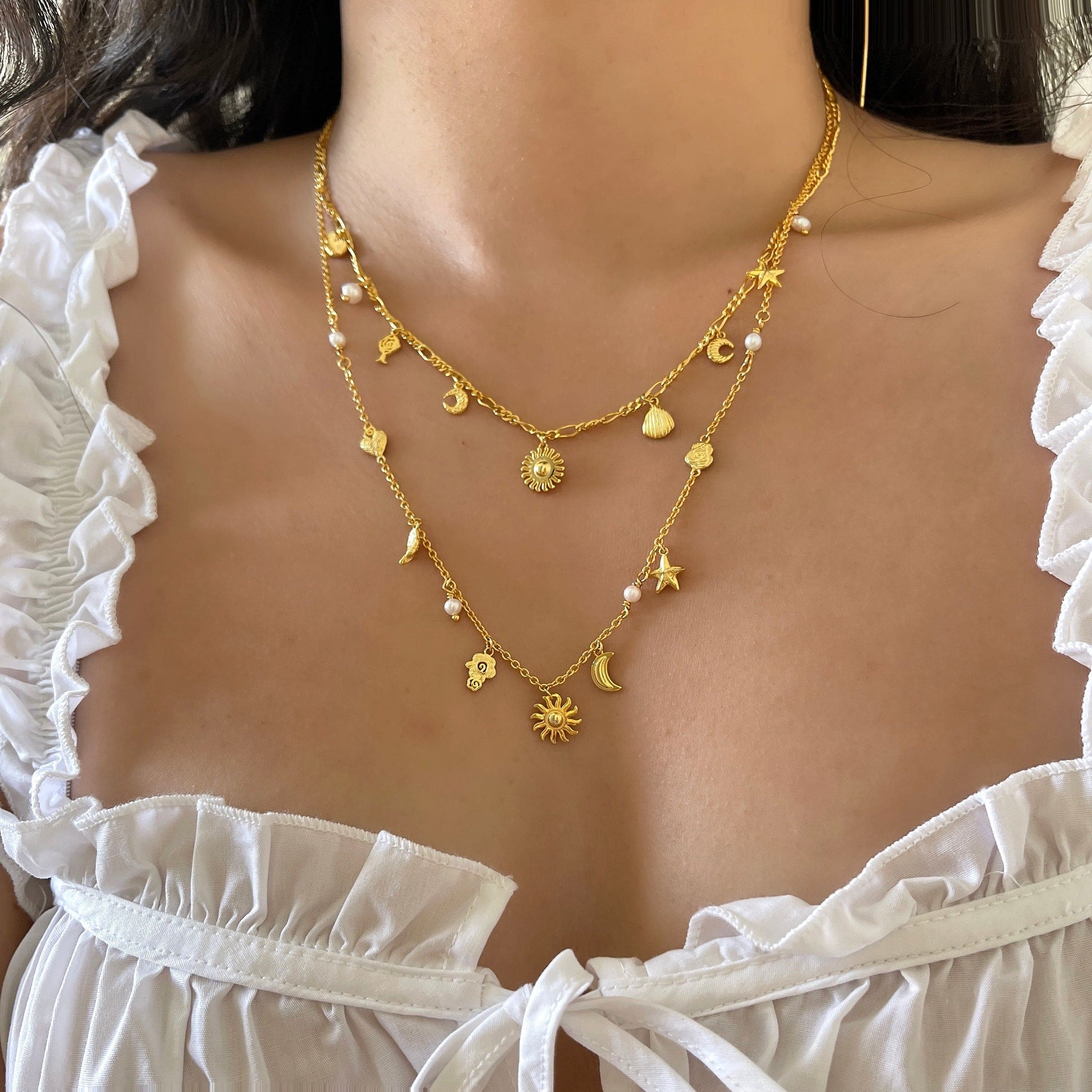 En kvinde bærer en hvid flæset top og to lag guldhalskæder, herunder Pallantis Smykkers Ocean Secret Halskæde, prydet med små sol-, månen-, stjerne- og andre vedhængsformer, der hviler lige over hendes bryst.