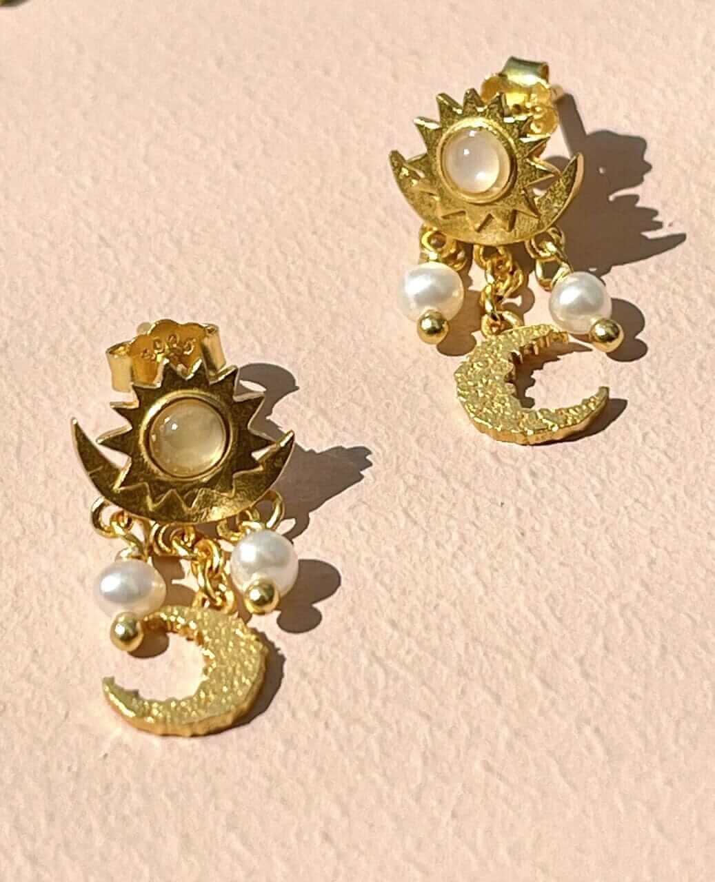 Pallantis Sol Øreringe Med Månesten har et sol- og måne-design med dinglende detaljer, inkl. to små perler og en struktureret guldmåne, på hvid baggrund—perfekte til at tilføje et himmelsk touch til dit look.