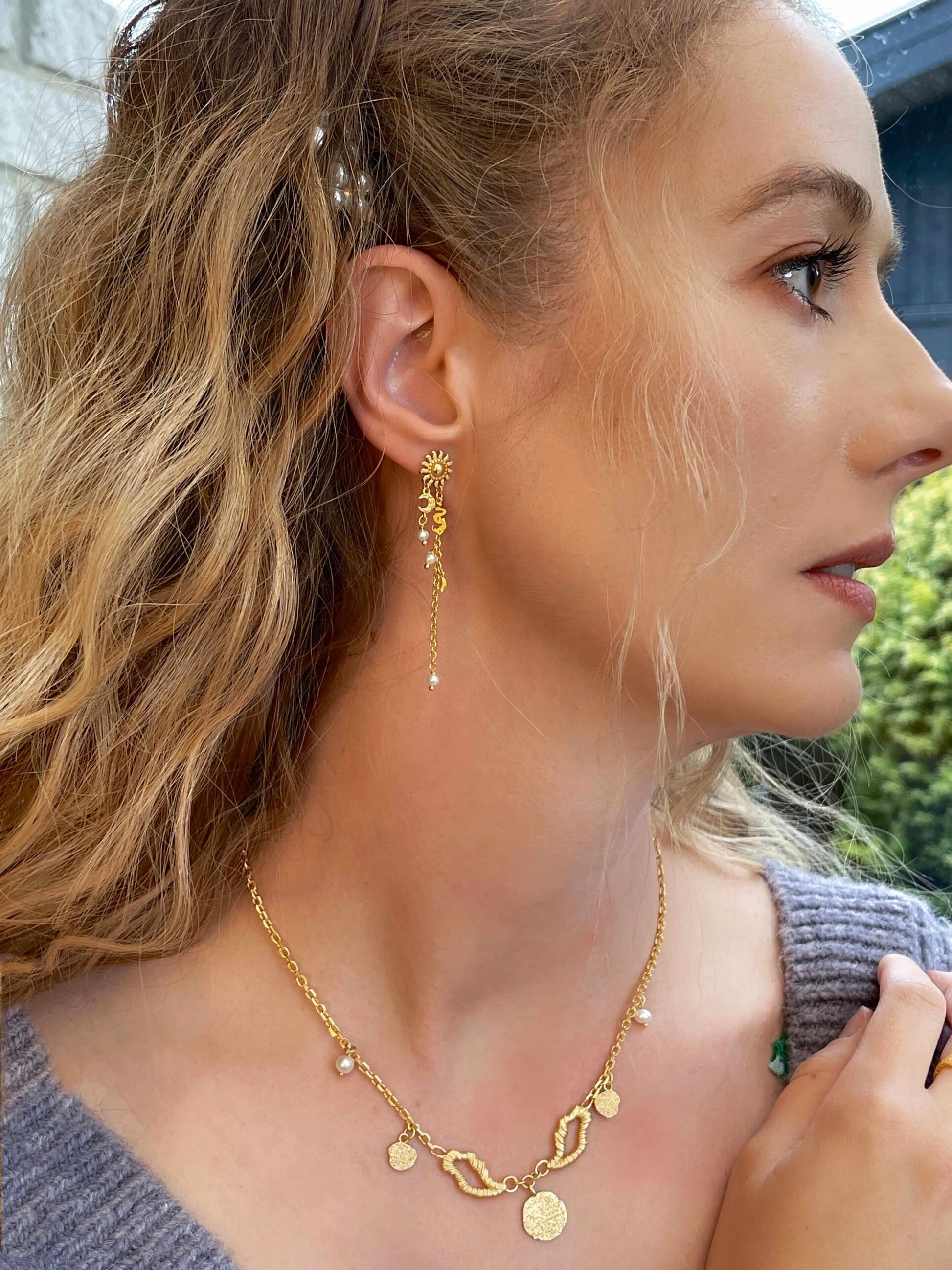 En kvinde med bølget blond hår bærer Pallantis Vinge Halskæde, prydet med møntvedhæng og perler, sammen med en dinglende guldørering med sol og blomster. Hun stirrer til højre, stående udendørs midt i frodigt grønt.