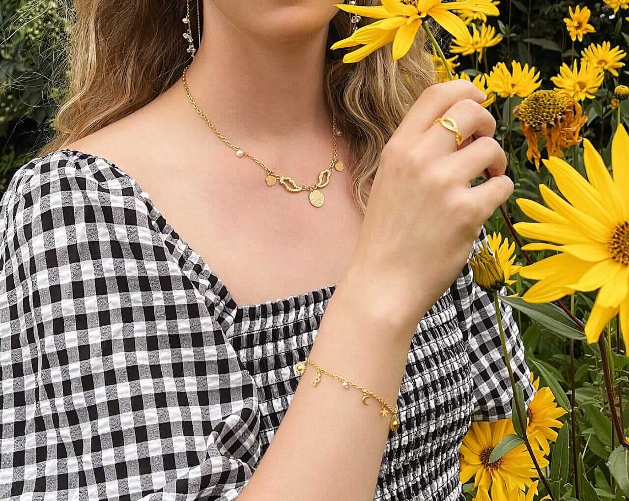 En kvinde i en sort-hvid ternet kjole holder en gul blomst og bærer Pallantis-guldsmykker, herunder Vinge Halskæde, armbånd og ring, med gule blomster, der blomstrer i baggrunden.